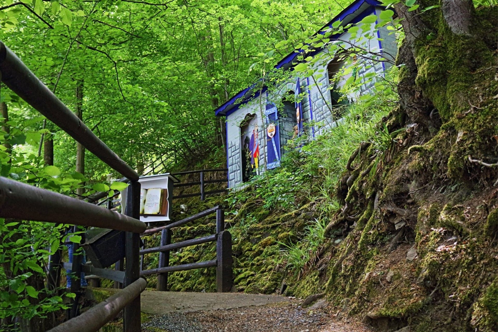 Waldweg mit Holzgeländer, darüber am Hang ein Märchenhaus mit Erzähltafel